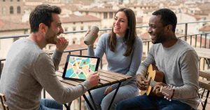 A group of people from different cultures singing along with one holding a guitar and the other has a laptop showing a sing-along platform on the screen